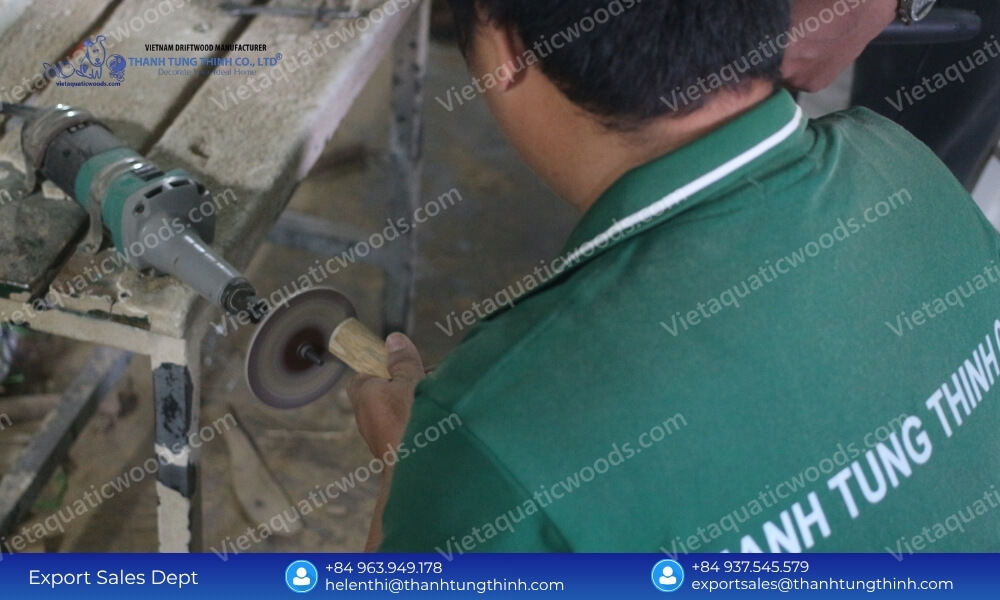 A factory artisan in a green shirt utilizing a mechanical grinding wheel to sand a coffee wood dog chew vietnam stick